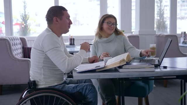 individual teaching for invalid, educator woman into glasses conducts training for disabled men on wheelchair using smart computer technology and books in restaurant