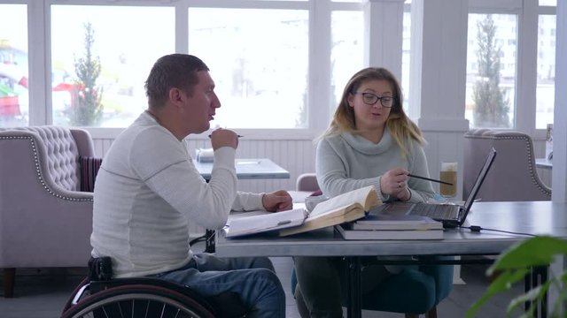 individual training for disabled, smart teacher female into glasses provides teaching for invalid man on wheelchair using modern computer technology and books in cafe