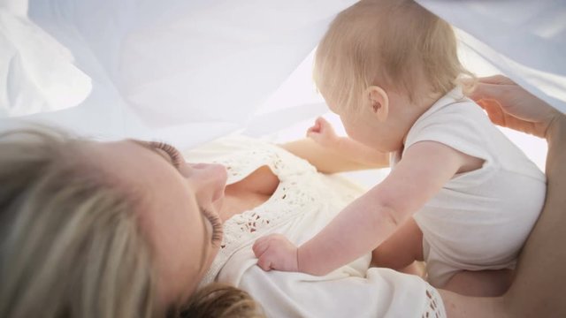 Cute baby boy chilling with loving mother under the white scarf in the park 