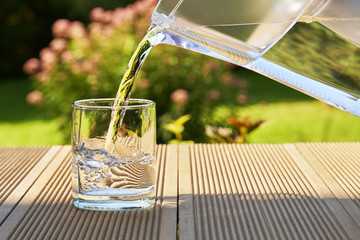 Pouring clear filtered water from a water filtration jug into a glass in green summer garden in a...