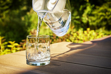 Pouring clear filtered water from a water filtration jug into a glass in green summer garden in a...