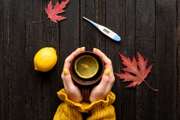 Mug of tea in female hands. Lemon and thermometer. Cold treatment. Wooden background. Top view