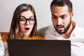 Portrait of surprised contemporary couple sitting on the yellow sofa in the light living room and looking on the fantastic proposition in the online shop on the laptop, modern technology