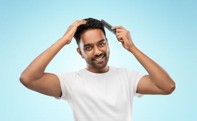 Fototapeta premium grooming, hairstyling and people concept - smiling young indian man brushing hair with comb over blue background