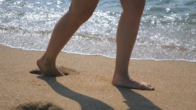 Close up of female feet walking on golden sand at the beach with ocean waves at background. Legs of young woman stepping at sand. Barefoot girl at the sea shore. Summer vacation holiday. Slow motion