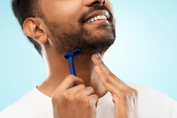 grooming and people concept - close up of young indian man shaving beard with manual razor blade...