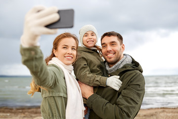 family, leisure and people concept - happy mother, father and little daughter taking selfie by smartphone on autumn beach