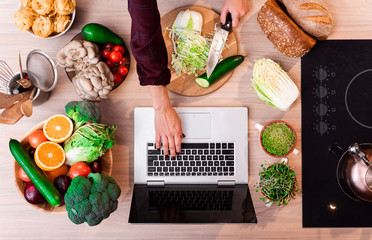 women in the kitchen searching for recipes on her laptop with food ingredients and fresh vegetables on a wooden table. seedlings.  top view. flat lay