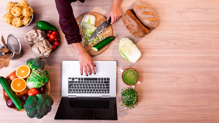 women in the kitchen searching for recipes on her laptop with food ingredients and fresh vegetables  on a wooden table on the left. top view. flat lay. space