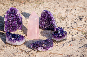 1 rose quartz gemstone surround by 5 amethyst crystals laying on the beach and in the grass