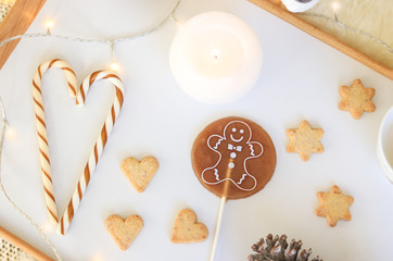 Top view of Christmas sweets on white tray. Candy cane, round gingerbread man lollipop, biscuits in heart and star shape, candle and garland lights. Flat lay. Cozy Christmas concept