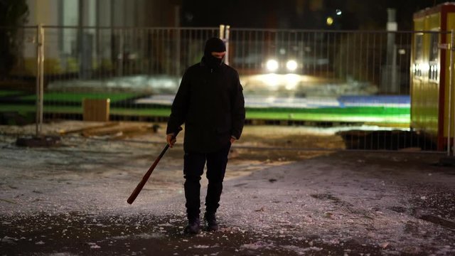 A man dressed in dark clothes and a balaclava goes with a bat in an abandoned park. There is an early winter outside and garlands hang around. His face is not visible in backlight. He is set up to fig