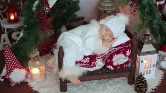 Adorable little toddler baby boy dressed in canta claus costume, sleeping in baby bed in front of teepee decorated for Christmas