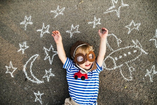 Portrait of smiling toddler wearing pilot hat and goggles lying on asphalt painted with airplane, moon and stars