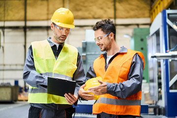 Two men wearing protective workwear holding clipboard and talking in factory