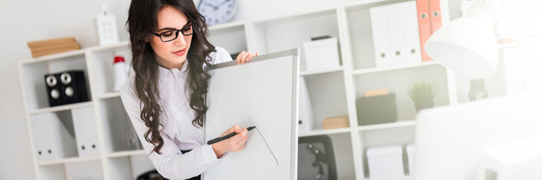 A beautiful young girl stands near an office desk and draws a magnetic marker on the magnetic board.