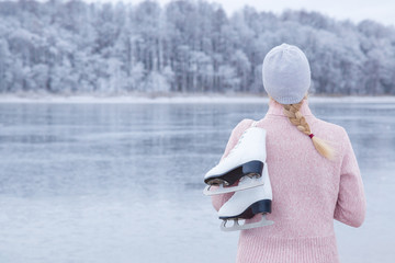 Young blond woman in pink sweater and hat staring at ice of lake and holding white skates over shoulder in freezing winter day. Back view of ice skater. Outdoor activities on weekends in cold weather. © fotoduets