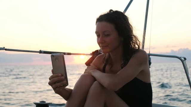Young woman is sailing yacht and uses a smartphone - takes a selfie at sunset
