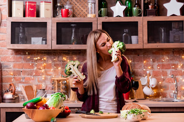 Young girl with blonde long hair of European appearance in a plaid shirt at the loft kitchen is bites broccoli. Assorted vegetables in a wooden plate on a light wooden table. close up