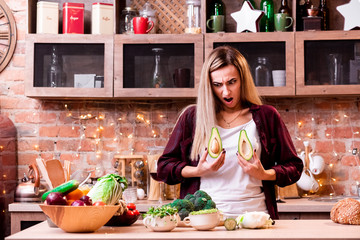 Happy Blonde girl with a surprised face in a white shirt holds avocado halves on her breast levels and makes grimaces in the loft kitchen. Concept: proper nutrition - is a guarantee of health.