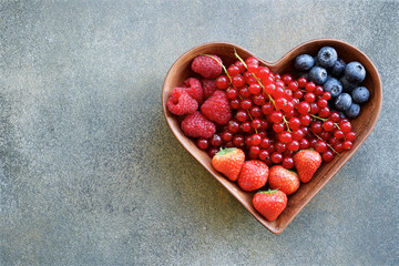 Heart shaped plate of raspberries, strawberries, red currants and blueberries.  Concrete texture with shades of green, brown and gray colors