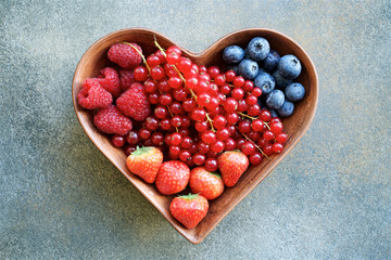 Heart shaped plate of raspberries, strawberries, red currants and blueberries.  Concrete texture with shades of green, brown and gray colors