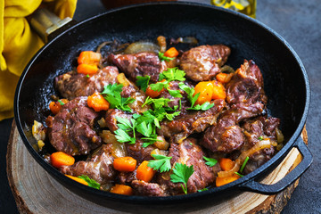 Pork cheeks stewed with vegetables in an iron pan, sliced bread, olive oil, with a dark background in a single shot above. - obrazy, fototapety, plakaty