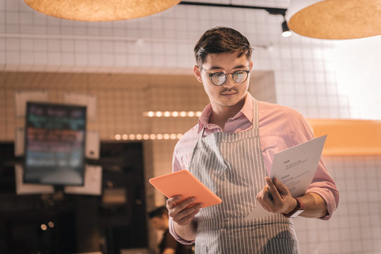 Male entrepreneur wearing striped apron holding menu of his restaurant
