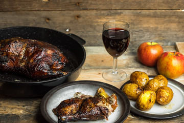goose baked with apples and potatoes, next to a glass of red wine on a wooden background, rustic table