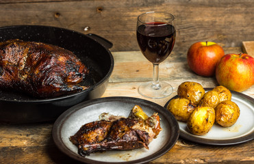 goose baked with apples and potatoes, next to a glass of red wine on a wooden background, rustic table