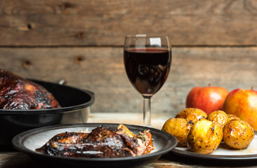 goose baked with apples and potatoes, next to a glass of red wine on a wooden background, rustic table