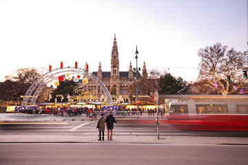 Vienna Rathausplatz and town hall at Christmas. People visiting the Christkindlmarkt, therefore waiting at traffic light to cross the road as blurred tram is passing by.