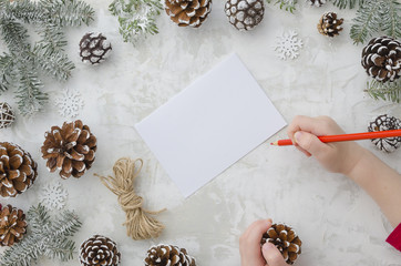 Child writes the letter to Santa Claus. Child's hands, the sheet of paper, red pencil and Christmas decorations pine cones and fir tree background. Concept of New year and Christmas. Top view