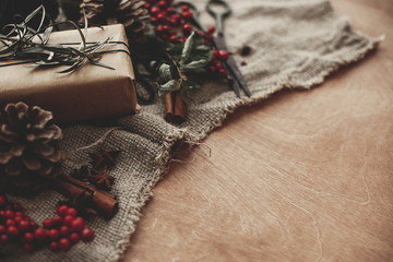 Stylish rustic christmas gift box with green branch and fir branches, red berries, pine cones, cinnamon on rustic wooden background. Simple eco friendly present wrapping. Atmospheric mood