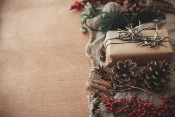 Fototapeta premium Stylish rustic christmas gift box with green branch and fir branches, berries, anise, pine cones, cinnamon on rustic wooden background. Simple eco present. Atmospheric mood. Space for text