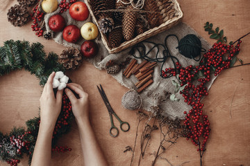 Rustic Christmas wreath flat lay. Hands putting cotton on fir branches, red berries and pine cones,thread, scissors, cinnamon on rustic wooden background. Making wreath at holiday workshop