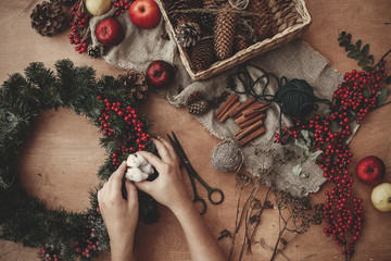 Rustic Christmas wreath flat lay. Hands putting cotton on fir branches, red berries and pine cones,thread, scissors, cinnamon on rustic wooden background. Making wreath at holiday workshop