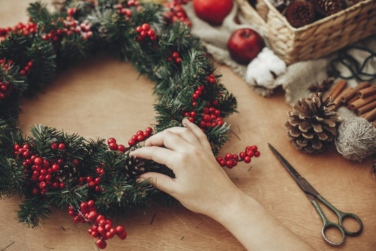 Hands making rustic christmas wreath with fir branches, red berries,pine cones,rope, scissors, cinnamon, cotton on rustic wooden background. Atmospheric moody image at holiday workshop