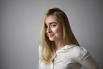 Cheerful mysterious young Europeanfemale with long blonde hair looking at camera with playful smile, having some secrets, being in good mood, posing isolated against blank gray wall background