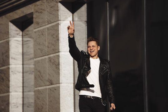 People, urban lifestyle and body language concept. Horizontal image of joyful handsome European guy in leather jacket making sign with raised hand, hailing cab, standing outside modern building