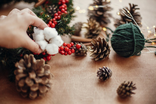 Hand putting cotton on wreath,pine cones, fir branches, red berries, thread, cinnamon,  lights on rustic wooden background. Details for making christmas wreath at workshop. Atmospheric image