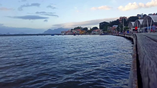 Scene of the Santa Maria river in the late afternoon. View of the deck in the Caieiras Island (Ilha das Caieiras), Vit&oacute;ria-ES. Houses, boats and people in the background. Clouded sky.