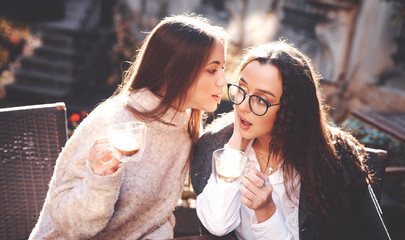 two beautiful stylish women sitting at the table in street cafe, drinking coffee and talking. women having lunch at bright sunny autumn day and gossiping, discussing something