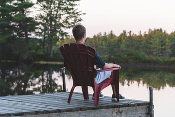 Fototapeta premium Young man relaxing on an Adirondack chair and looking at a calm river at sunset. Muskoka, Ontario, Canada.