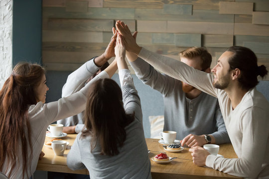 Happy multiethnic friends meeting in cafe giving high five showing support and unity, excited young people sitting in coffeeshop enjoying time together raising hands joining, promising friendship