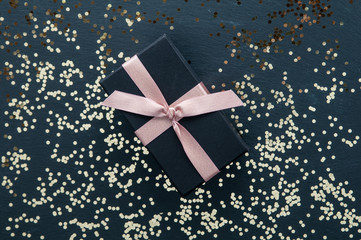 Gift in a black box tied with a pink ribbon on black slate background with gold glitter