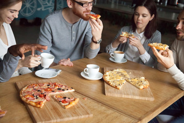Multiethnic friends have fun in Italian restaurant, eating pizza and drinking coffee, diverse colleagues talk at cafe table, enjoy tasty food, young people speak laughing hanging out in pizzeria