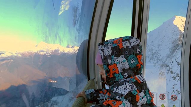 Little Girl Looking Out From Window Inside Cable Car Moving On Snowy Slope In Winter Mountain. Close Up Baby Girl Riding Up In Ski Elevator On Snowy Mountain On Ski Resort