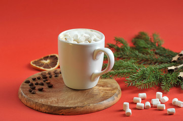 Cappuccino with marshmallows in a white cup. The composition is supplemented with a spruce branch, decorative wooden stars and coffee beans. Red background. Close-up. 