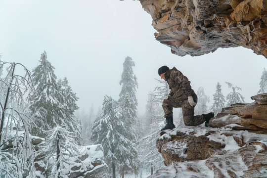 Man Hiker Looking Down From High Altitude Standing On Scenic Rock, Winter Mountains And Fir Forest Covered With Snow At Background.